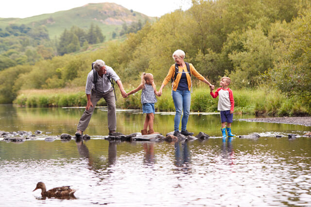 Two young children holding hands with their grandparents walking across a river on stepping stones