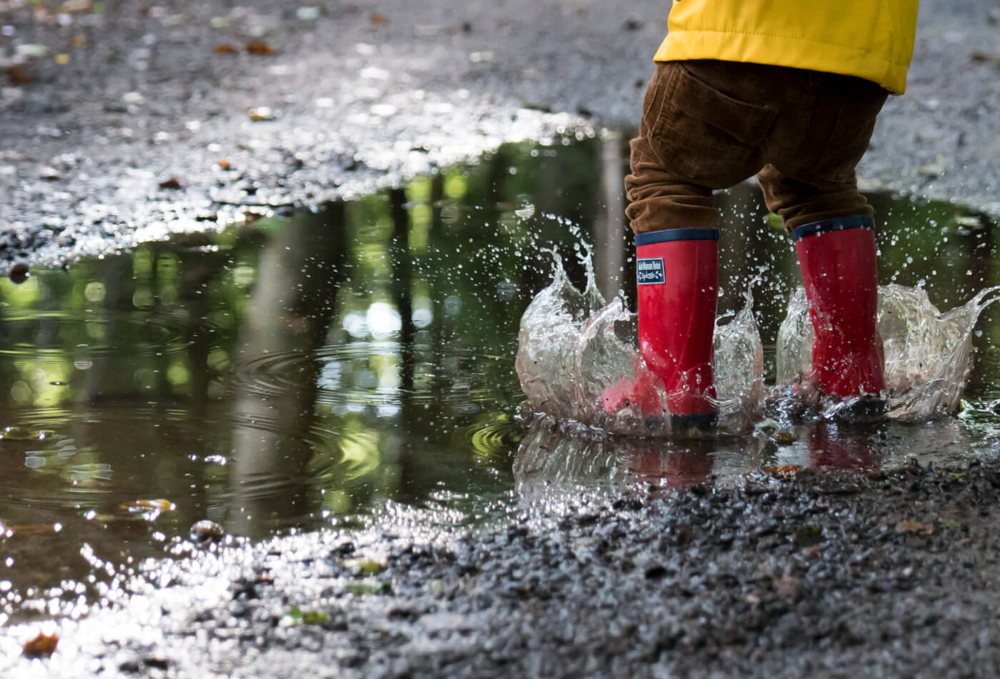 A child wearing red wellington boots and a yellow coat jumping into a puddle