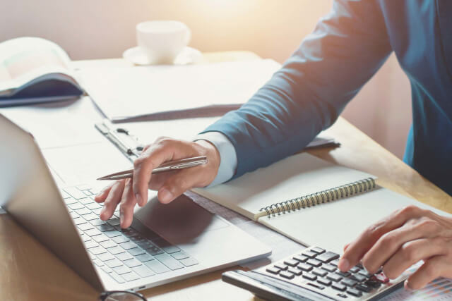 A man sat at a desk typing on a laptop and a calculator