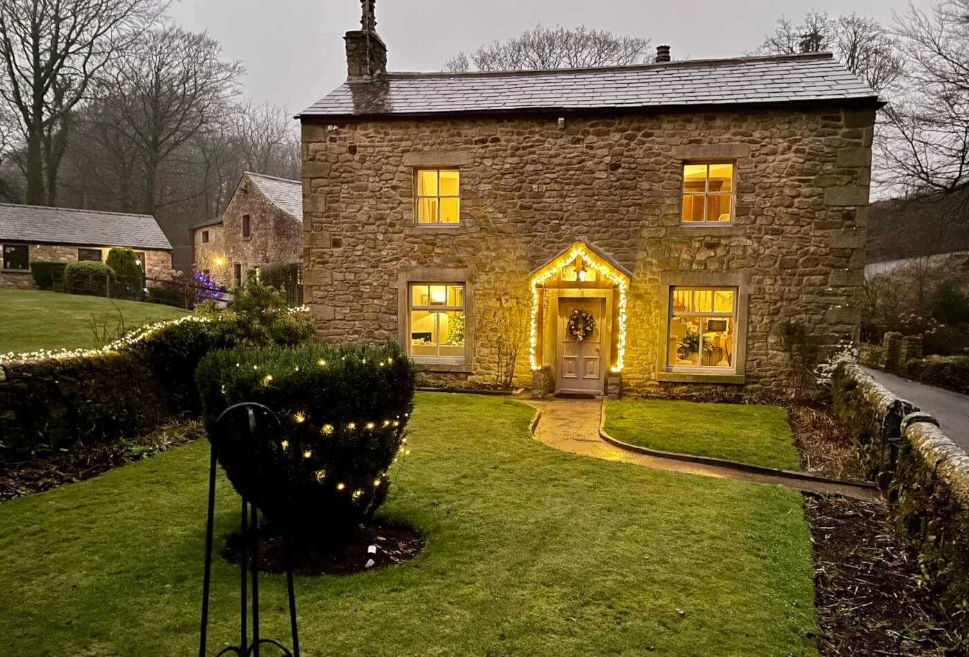 A stonebuilt cottage with lights around the doorframe and on a tree in the green front garden