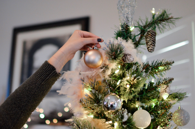 Person holding a beige coloured bauble next to a decorated Christmas tree. 