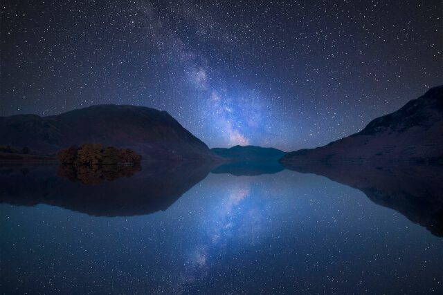 A view across a lake to the mountains beyond with a dark sky decorated in bright colour