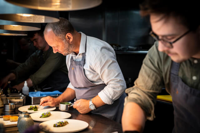 Simon Rogan serving three plates of food in the restaurant of a kitchen