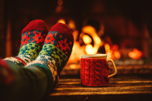 Person with festive socks and feet up on a table with a festive mug in front of a fire.