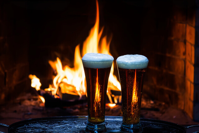 Two pints of beer on a table in front of a roaring open fire