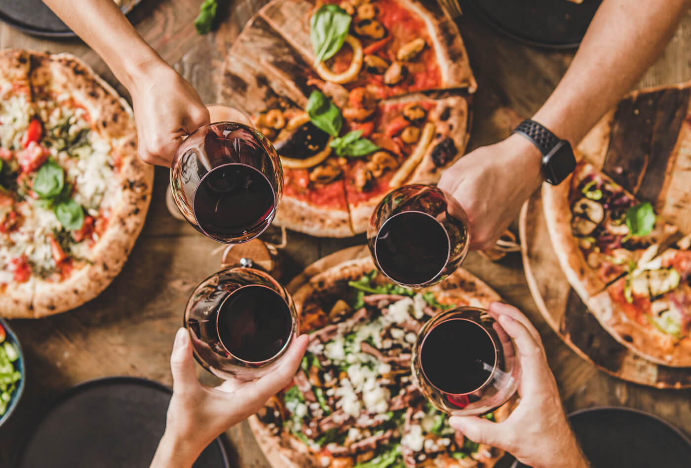 People clinking glasses of red wine above a table of pizzas