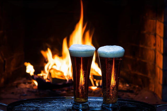 Two pints of beer on a table in front of an open fire 